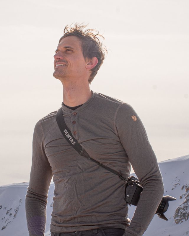 Ryan Lichtenwalter smiling on a snowy mountain ridge, a camera on a strap across his chest